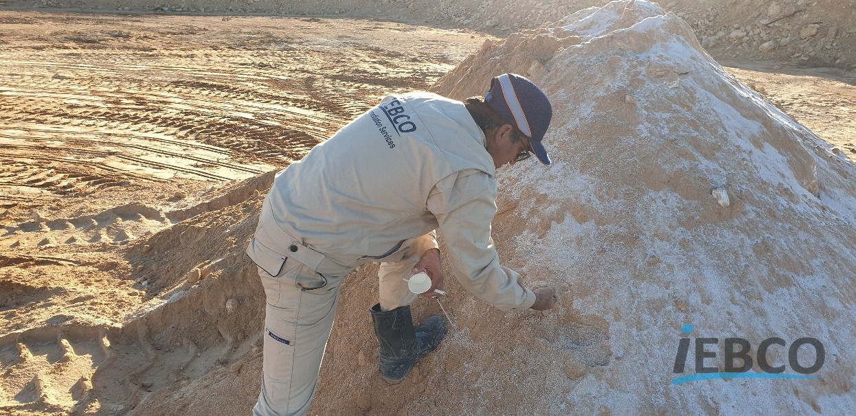 کارشناس IEBCO خم‌شده روی توده مواد در محوطه پروژه، با ظرف نمونه‌برداری در دست.